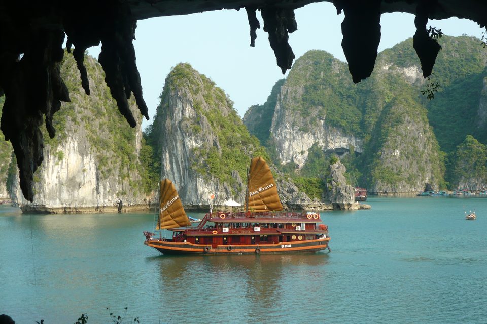 A traditional junk boat with ochre sails on Ha Long Bay, framed by limestone karsts and the mouth of a sea cave.