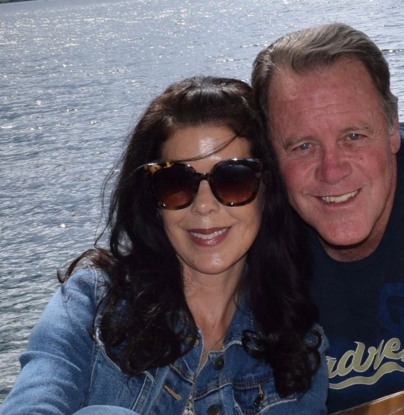 Therese and Steve Devan side by side on a lake shore, mountains behind them.